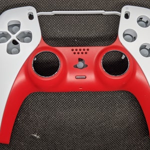 May include: A two-tone red and white game controller shell. The controller has a red centre section with a PlayStation logo and a white top and side sections. The controller has grey buttons and a grey connecting bar.