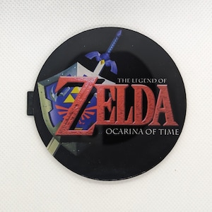 May include: A round, black button featuring the text "THE LEGEND OF ZELDA OCARINA OF TIME" in red and white. The button also displays a shield, sword, and the Zelda logo in blue and gold.