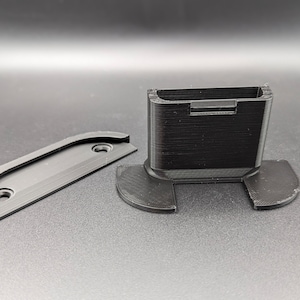 Puede incluir: Componentes de plástico negro impresos en 3D, que incluyen un soporte rectangular con una ranura y una placa curva con dos agujeros. Las piezas están diseñadas para encajar. Los artículos están sobre una superficie oscura.