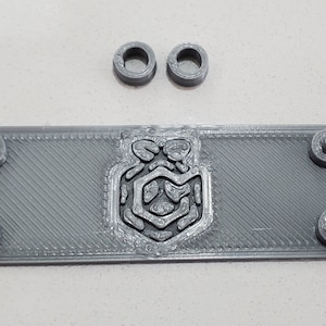May include: A gray 3D-printed rectangular component with a detailed emblem in the center and two small circular pieces above it. The component has rounded corners, screw posts, and a loop on each end.
