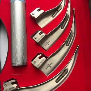 May include: A medical instrument set in a red velvet-lined case. The set includes a silver-colored laryngoscope handle and several curved blades. The handle is cylindrical with a textured grip. The blades are marked with "BMD 712" and "boromed".