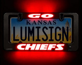May include: A black rectangular license plate frame with integrated LED lighting. The top bar glows red with the word GO, and the bottom bar glows red with the word CHIEFS. The frame surrounds a Kansas license plate featuring a building and wheat.