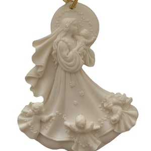 Puede incluir: Adorno de color marfil que representa a una mujer sosteniendo a un bebé, rodeada de ángeles. La mujer lleva un vestido fluido y un halo. El adorno está suspendido por una cinta dorada. Detalles florales decorativos.