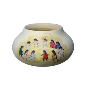 Puede incluir: Una maceta de cerámica blanca con una escena pintada a mano de niños que se toman de la mano y bailan en círculo. Los niños visten ropa colorida y el fondo es un cielo de tono amarillo. La maceta está firmada "De Gregoria" en la parte inferior.