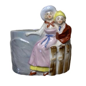 May include: A vintage ceramic planter depicting a couple in period attire. The figures are painted in pastel shades, with the woman in a pink dress and the man in a brown jacket. The planter has a pale blue exterior and a rustic fence detail.