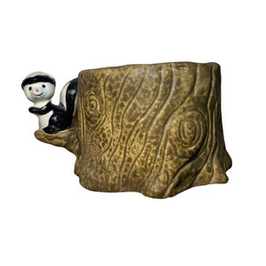 May include: A ceramic planter shaped like a tree stump, with a skunk figurine on the side. The planter is brown with a textured bark design. The skunk is black and white with a smiling face and a black hat.
