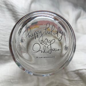 Puede incluir: Vaso de vidrio transparente con las palabras "happy holidays 2020" y la firma "Ondine Jarin" en escritura negra. Tambi&eacute;n son visibles las palabras "for ANTHROPOLOG".
