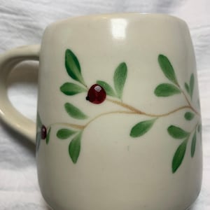 Puede incluir: Taza de cer&aacute;mica color crema con asa curva. La taza est&aacute; decorada con hojas verdes y bayas rojas pintadas sobre un tallo marr&oacute;n claro. La taza tiene un dise&ntilde;o simple y elegante.