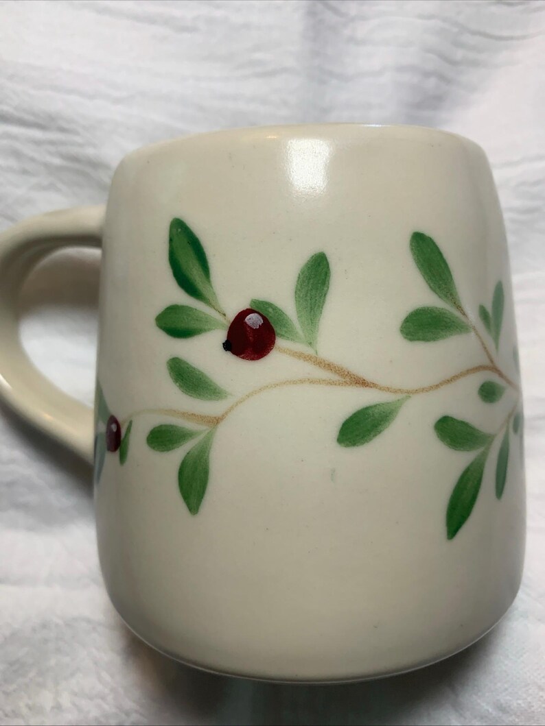 Puede incluir: Taza de cer&aacute;mica color crema con asa curva. La taza est&aacute; decorada con hojas verdes y bayas rojas pintadas sobre un tallo marr&oacute;n claro. La taza tiene un dise&ntilde;o simple y elegante.
