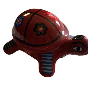 Könnte beinhalten: Eine rote Keramik-Schildkrötenfigur mit einem bemalten Panzer mit schwarzen und weißen Linien und bunten Blumen. Die Schildkröte hat blaue und weiße Streifen an den Beinen.