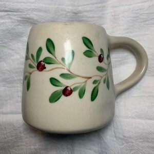 Puede incluir: Taza de cer&aacute;mica de color crema con asa curva. La taza est&aacute; decorada con hojas verdes y bayas rojas pintadas sobre una vid marr&oacute;n. La taza tiene una forma cuadrada redondeada.