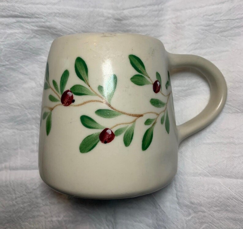 Puede incluir: Taza de cer&aacute;mica de color crema con asa curva. La taza est&aacute; decorada con hojas verdes y bayas rojas pintadas sobre una vid marr&oacute;n. La taza tiene una forma cuadrada redondeada.