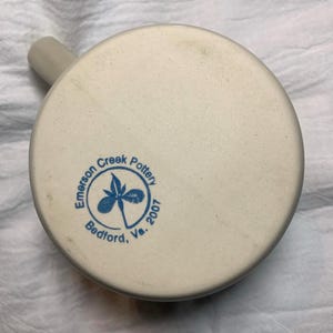 Puede incluir: Una taza de cer&aacute;mica de color crema con asa. La base de la taza presenta un logotipo circular azul con el texto "Emerson Creek Pottery Bedford, Va. 2007" y un dise&ntilde;o floral.