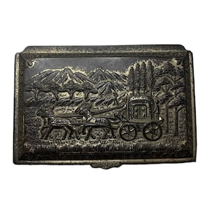 May include: A silver metal trinket box with an embossed design of a horse-drawn carriage traveling through a mountainous landscape.