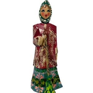 Può includere: Una statuetta in legno dipinta a mano raffigurante una persona che indossa un cappotto rosso con motivi bianchi e un foulard verde acqua. La figura tiene in mano un lungo e sottile bastone di legno e indossa una lunga gonna plissettata decorata con motivi floreali verdi, rosa e blu.