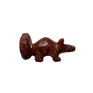 May include: A small, hand-carved, red and white speckled stone animal figurine. The animal is a stylized version of a badger with blue eyes.