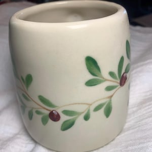 Puede incluir: Una taza de cer&aacute;mica de color crema con forma redondeada. La taza est&aacute; decorada con un dise&ntilde;o de hojas verdes y bayas rojas. El dise&ntilde;o est&aacute; pintado alrededor de la taza en un patr&oacute;n repetitivo. La taza tiene un acabado liso y brillante.