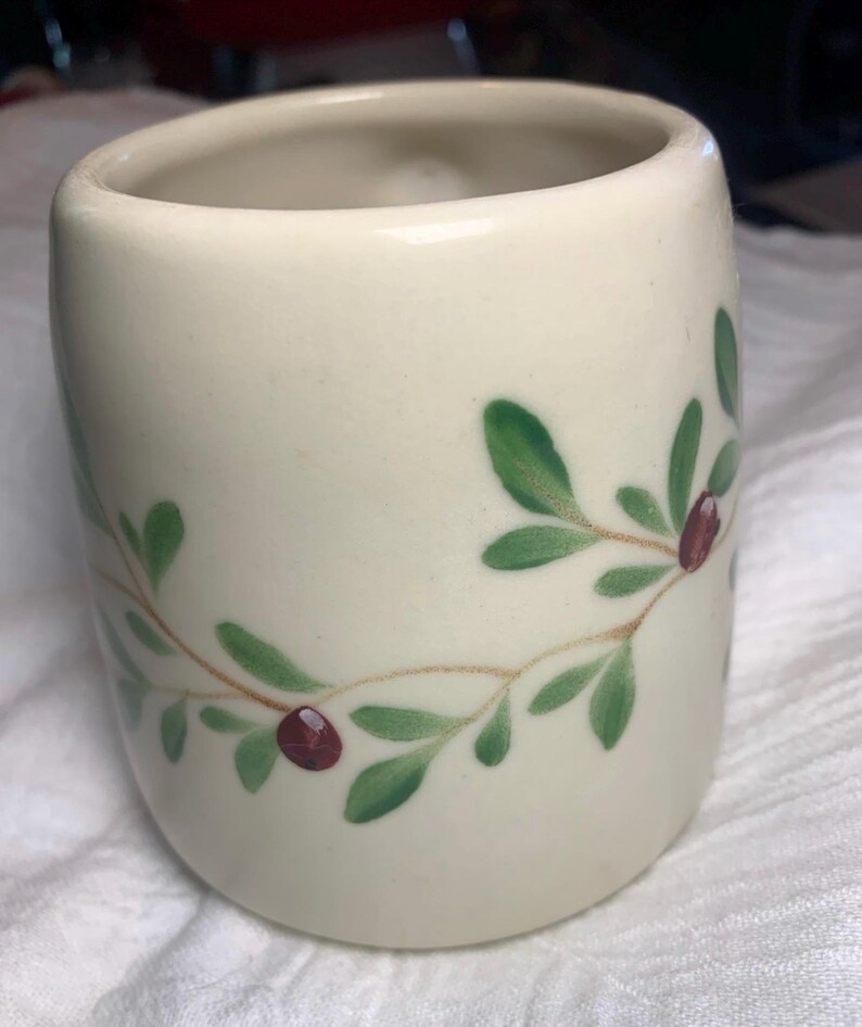 Puede incluir: Una taza de cer&aacute;mica de color crema con forma redondeada. La taza est&aacute; decorada con un dise&ntilde;o de hojas verdes y bayas rojas. El dise&ntilde;o est&aacute; pintado alrededor de la taza en un patr&oacute;n repetitivo. La taza tiene un acabado liso y brillante.