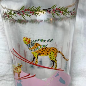 Puede incluir: Vaso transparente con borde dorado y dise&ntilde;o festivo. El vaso presenta un leopardo amarillo y naranja con bufanda esquiando por una monta&ntilde;a rosa. Acebo verde y rojo adorna la parte superior.