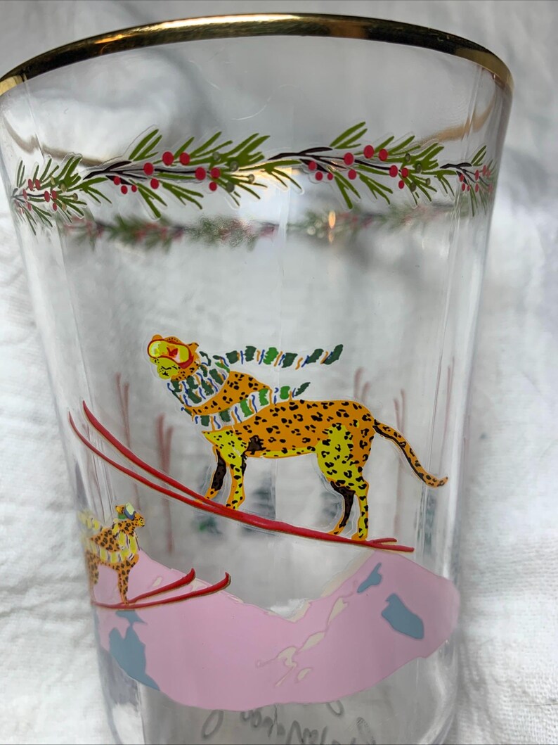 Puede incluir: Vaso transparente con borde dorado y dise&ntilde;o festivo. El vaso presenta un leopardo amarillo y naranja con bufanda esquiando por una monta&ntilde;a rosa. Acebo verde y rojo adorna la parte superior.