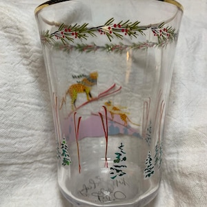 Puede incluir: Vaso de vidrio transparente con borde dorado y dise&ntilde;o de escena invernal. El vaso presenta un esquiador, &aacute;rboles y una guirnalda de hojas verdes y bayas rojas. La parte inferior del vaso tiene texto manuscrito y el a&ntilde;o 2016.