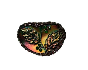 May include: An oval-shaped brooch with a leaf and berry design. The brooch features iridescent colors of green, gold, and purple. The leaves have detailed veining, and the berries are small and green. The edges are outlined with small, dark beads.