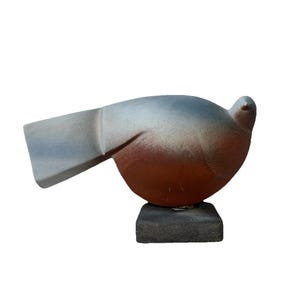 Könnte beinhalten: Keramikvogelskulptur in Grau- und Rottönen. Der Vogel sitzt auf einem kleinen, quadratischen, dunkelgrauen Sockel. Die Skulptur hat einen glatten, abgerundeten Körper und einen stilisierten Flügel. Ca. 15 cm lang.
