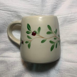 Puede incluir: Taza de cer&aacute;mica color crema con asa curva. La taza est&aacute; decorada con un dise&ntilde;o pintado a mano de hojas verdes y bayas rojas sobre una rama marr&oacute;n. La taza es de tama&ntilde;o est&aacute;ndar.