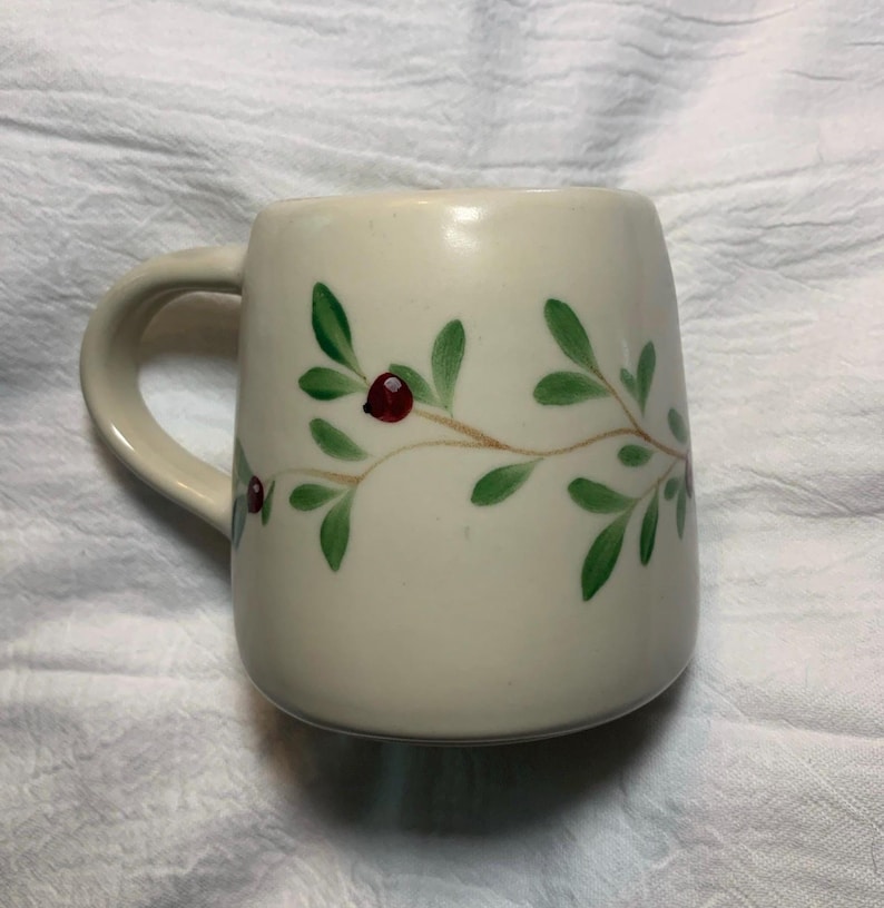 Puede incluir: Taza de cer&aacute;mica color crema con asa curva. La taza est&aacute; decorada con un dise&ntilde;o pintado a mano de hojas verdes y bayas rojas sobre una rama marr&oacute;n. La taza es de tama&ntilde;o est&aacute;ndar.