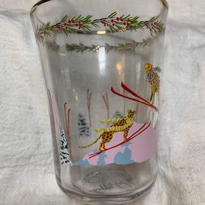 Puede incluir: Vaso de vidrio transparente con borde dorado y dise&ntilde;o de tem&aacute;tica invernal. El dise&ntilde;o presenta un esquiador, un leopardo sobre esqu&iacute;s y una guirnalda de hojas verdes y bayas rojas. El vaso mide aproximadamente 13 cm de alto.