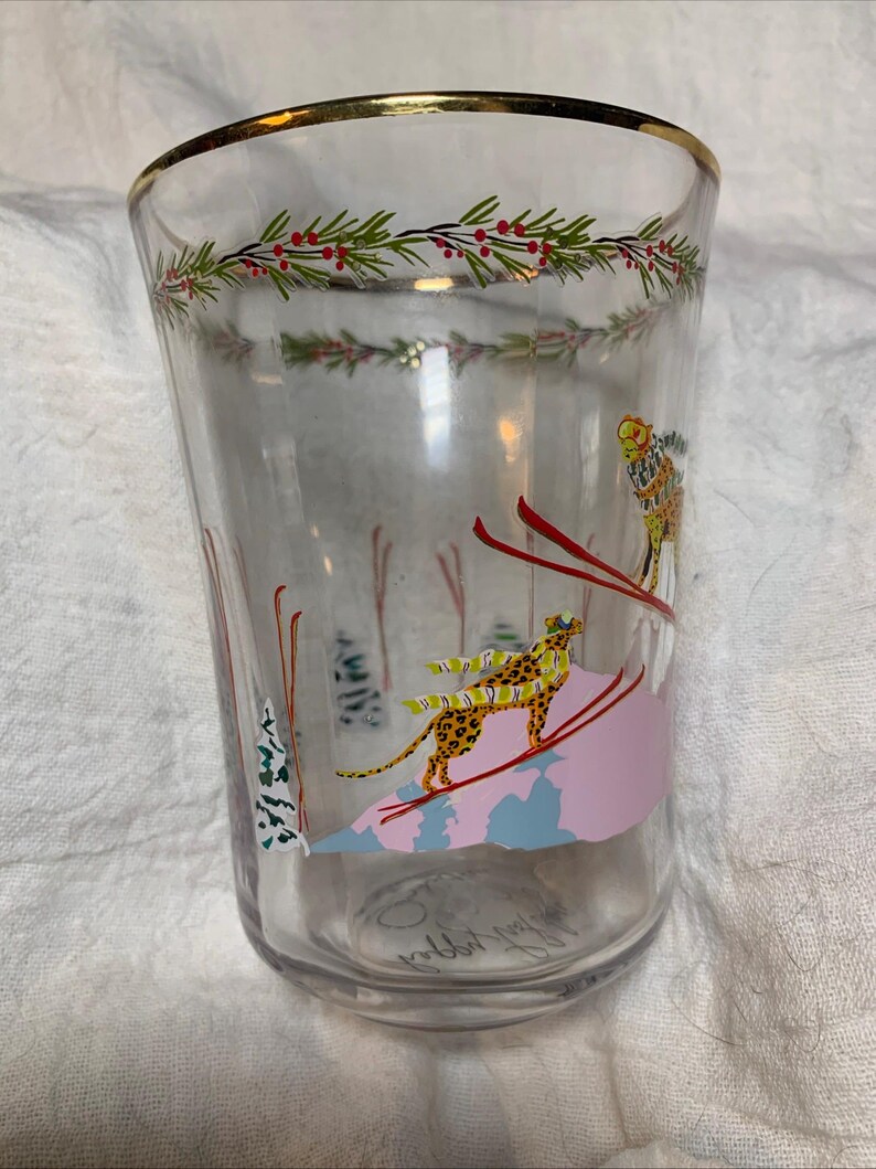 Puede incluir: Vaso de vidrio transparente con borde dorado y dise&ntilde;o de tem&aacute;tica invernal. El dise&ntilde;o presenta un esquiador, un leopardo sobre esqu&iacute;s y una guirnalda de hojas verdes y bayas rojas. El vaso mide aproximadamente 13 cm de alto.
