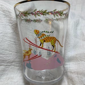 Puede incluir: Vaso de vidrio transparente con borde dorado y dise&ntilde;o festivo. El vaso presenta un guepardo esquiando por una monta&ntilde;a rosa, con un guepardo m&aacute;s peque&ntilde;o en el fondo. La parte superior del vaso tiene un dise&ntilde;o de acebo verde y rojo.