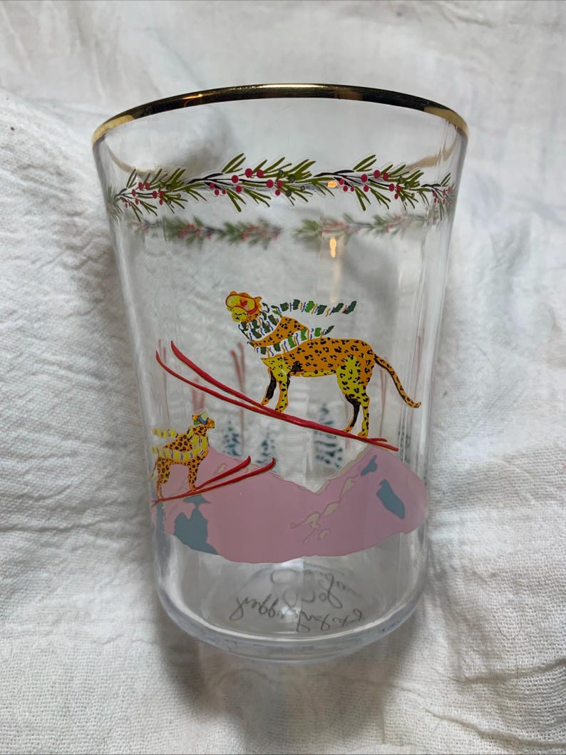 Puede incluir: Vaso de vidrio transparente con borde dorado y dise&ntilde;o festivo. El vaso presenta un guepardo esquiando por una monta&ntilde;a rosa, con un guepardo m&aacute;s peque&ntilde;o en el fondo. La parte superior del vaso tiene un dise&ntilde;o de acebo verde y rojo.