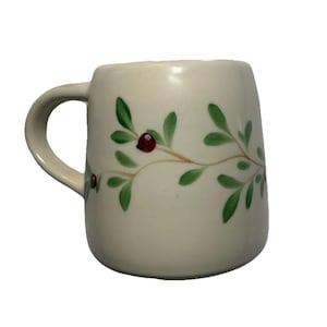 May include: Cream-colored ceramic mug with a curved handle. The mug is decorated with a painted design of green leaves and red berries on a brown stem. The mug has a smooth, glossy finish.