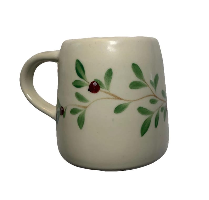 Puede incluir: Taza de cer&aacute;mica color crema con asa curva. La taza est&aacute; decorada con un dise&ntilde;o pintado de hojas verdes y bayas rojas sobre un tallo marr&oacute;n. La taza tiene un acabado liso y brillante.