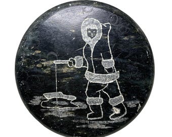Può includere: Una scultura rotonda in pietra nera e verde che raffigura una persona con un parka con cappuccio che pesca sul ghiaccio. La persona tiene una canna da pesca e c'è un buco nel ghiaccio.