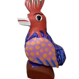 Puede incluir: Una figurita de pájaro de madera pintada a mano con cabeza roja, cuerpo azul y morado y lunares naranja. El pájaro está de pie sobre una base marrón.