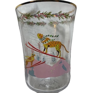 Puede incluir: Vaso de vidrio transparente con borde dorado y dise&ntilde;o de tem&aacute;tica invernal. El vaso presenta un leopardo esquiando por una monta&ntilde;a rosa, con un leopardo m&aacute;s peque&ntilde;o en el fondo. El dise&ntilde;o incluye ramas de hoja perenne y bayas rojas en la parte superior.