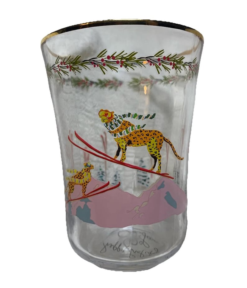 Puede incluir: Vaso de vidrio transparente con borde dorado y dise&ntilde;o de tem&aacute;tica invernal. El vaso presenta un leopardo esquiando por una monta&ntilde;a rosa, con un leopardo m&aacute;s peque&ntilde;o en el fondo. El dise&ntilde;o incluye ramas de hoja perenne y bayas rojas en la parte superior.