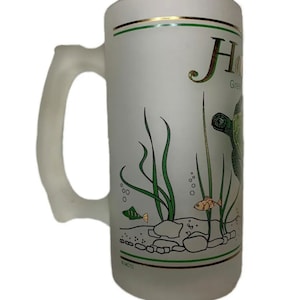Puede incluir: Una jarra de cerveza de vidrio esmerilado con un diseño verde y dorado que presenta una tortuga marina, algas marinas y peces. La jarra tiene el texto "Hawaii Green Sea Turtle" en la parte delantera.