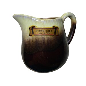 May include: A brown and yellow ceramic creamer with a brown glaze and a yellow band around the top. The creamer has a handle and a label that reads "Stonefield, Cassville, Wis."