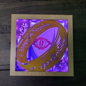 Op de afbeelding: Een ingelijst, verlicht kunstwerk met een gouden ring met de inscriptie "Een Ring om ze te regeren, Een Ring om ze te vinden, Een Ring om ze allen te brengen en in de duisternis te binden" in Elvish schrift. De ring heeft een gloeiend rood oog in het midden. Het kunstwerk is verlicht met paarse en blauwe lichten.