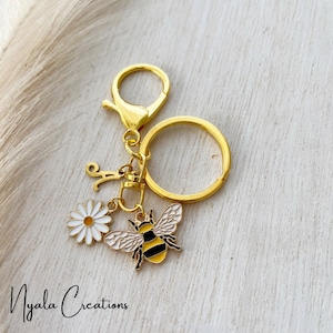 May include: Gold keyring with a bee charm, a white daisy charm, and a letter 'A' charm. The keyring is on a white wooden surface.