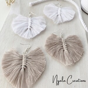 May include: Two pairs of handmade macrame earrings. One pair is white and the other pair is beige. The earrings are shaped like feathers and have a fringe detail.