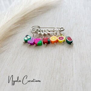 May include: A set of seven colourful fruit-shaped stitch markers attached to a silver safety pin. The markers include a green apple, a pink grape, a red strawberry, a red apple, a yellow lemon, a pink grapefruit, and a green watermelon.