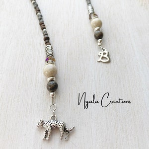 May include: Two beaded glasses chains with silver charms. One chain has a silver leopard charm and the other has a silver letter B charm. The chains are made with brown, white, and grey beads. The text "Nyala Creations" is visible in the image.