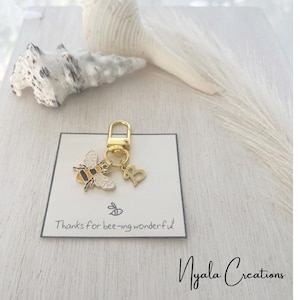 May include: A gold-coloured keyring with a bee charm and a letter "B" charm. The bee charm is black, white, and yellow. The keyring is attached to a white card with the text "Thanks for bee-ing wonderful!"