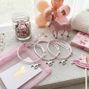 May include: Three light pink cord bracelets with silver-toned horse charms, a pink organza bag, a white card with a gold horse silhouette, a pink horse figurine with a party hat, a jar of star confetti, and napkins with horse and crown prints.