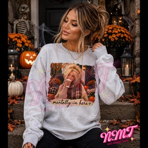May include: A light gray crewneck sweatshirt featuring a rectangular graphic print of a person holding a coffee cup in an autumn setting. The text "mentally i'm here" is printed in a cursive script below the image on the front of the garment.