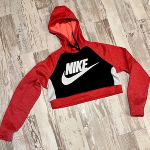 May include: Red and black cropped hoodie with the Nike logo in white. The hoodie has a hood and long sleeves. The body of the hoodie is black with the Nike logo, and the sleeves and hood are red.